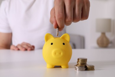 Photo of Man putting coin into yellow piggy bank at white table, closeup