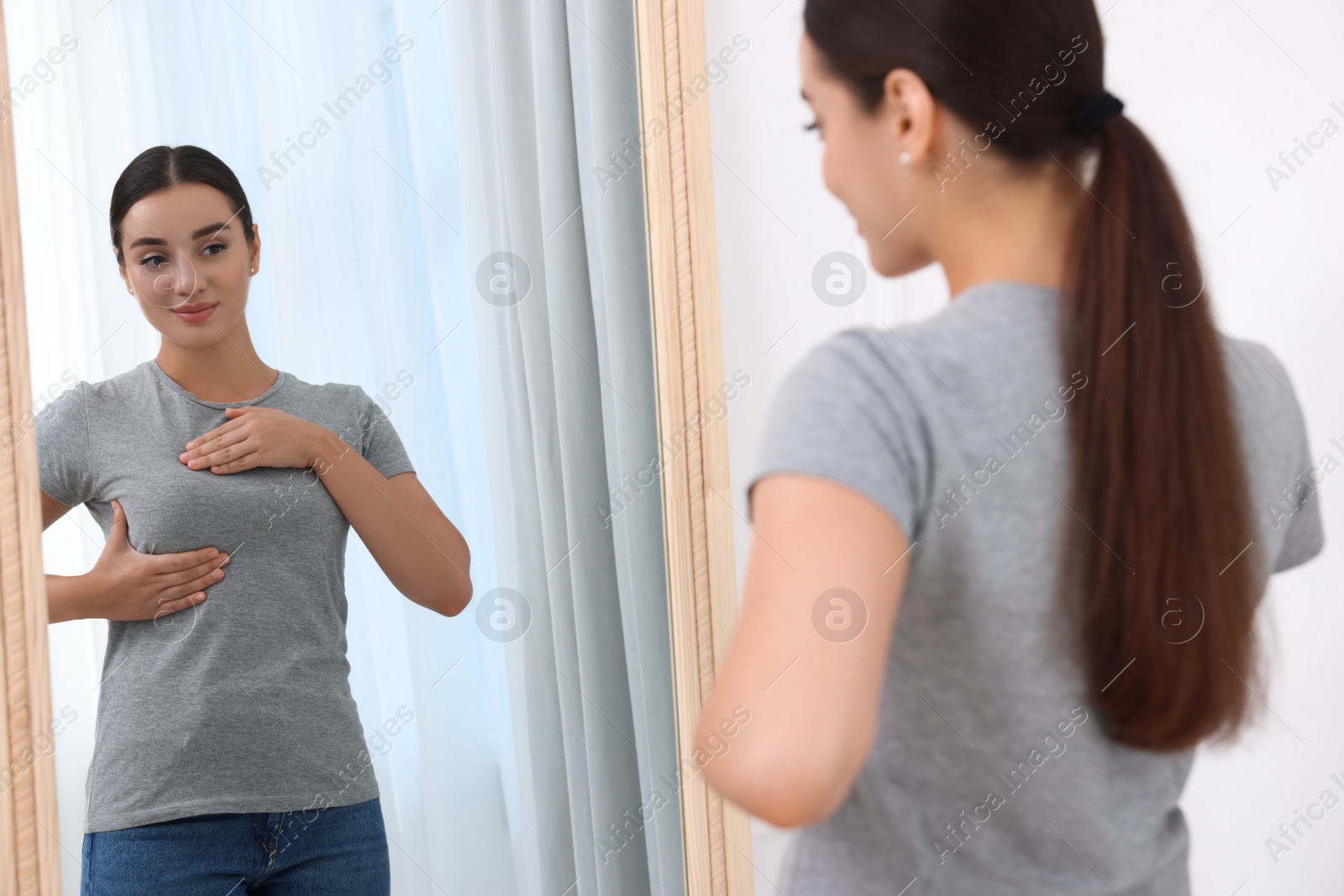 Photo of Beautiful young woman doing breast self-examination near mirror indoors