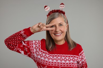 Happy senior woman in Christmas sweater and Santa headband on grey background