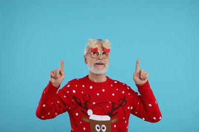 Senior man in Christmas sweater and funny glasses pointing at something on light blue background. Space for text