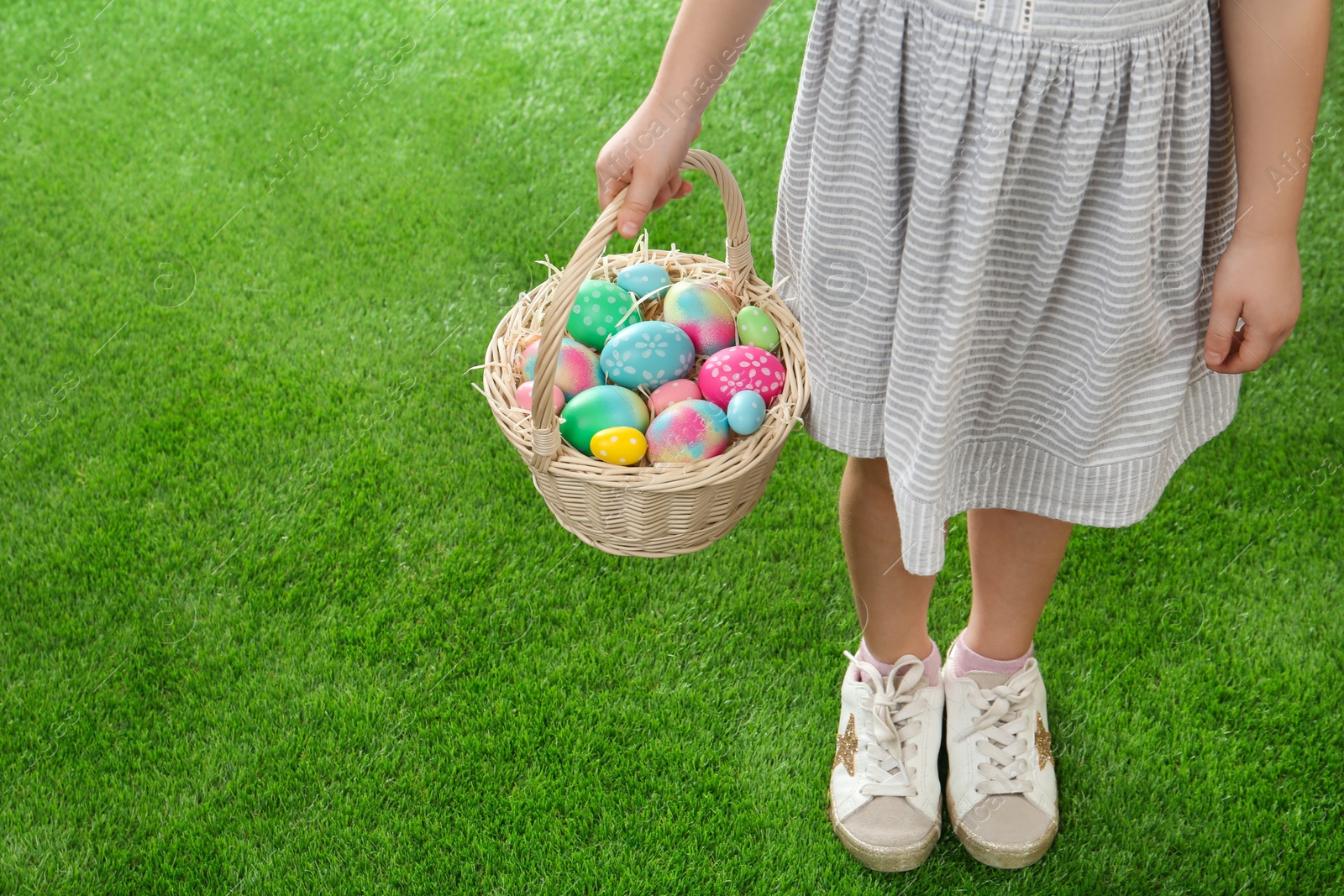 Photo of Little girl with basket full of Easter eggs on green grass, closeup. Space for text