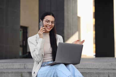 Happy young woman using modern laptop and talking on smartphone outdoors