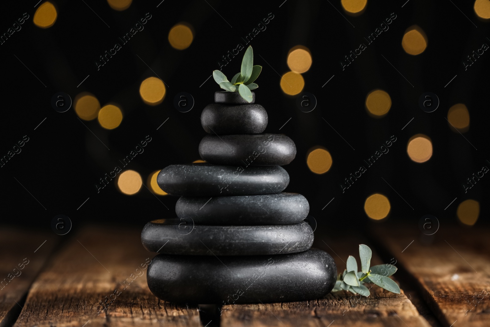 Photo of Stack of black spa stones with green branches on wooden table against blurred lights
