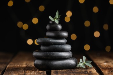 Stack of black spa stones with green branches on wooden table against blurred lights