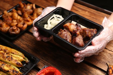 Photo of Waiter with plastic container of tasty shish kebab at wooden table, closeup. Food delivery service