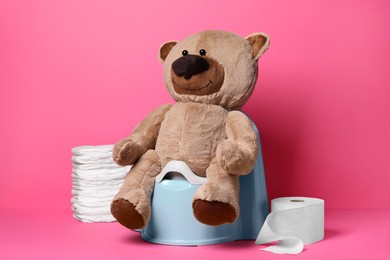 Photo of Teddy bear on baby potty, toilet paper and stack of diapers on pink background