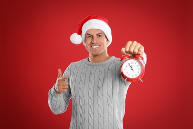 Photo of Man in Santa hat with alarm clock on red background. New Year countdown
