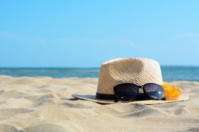 Hat with beautiful sunglasses and flowers on sand near sea. Space for text