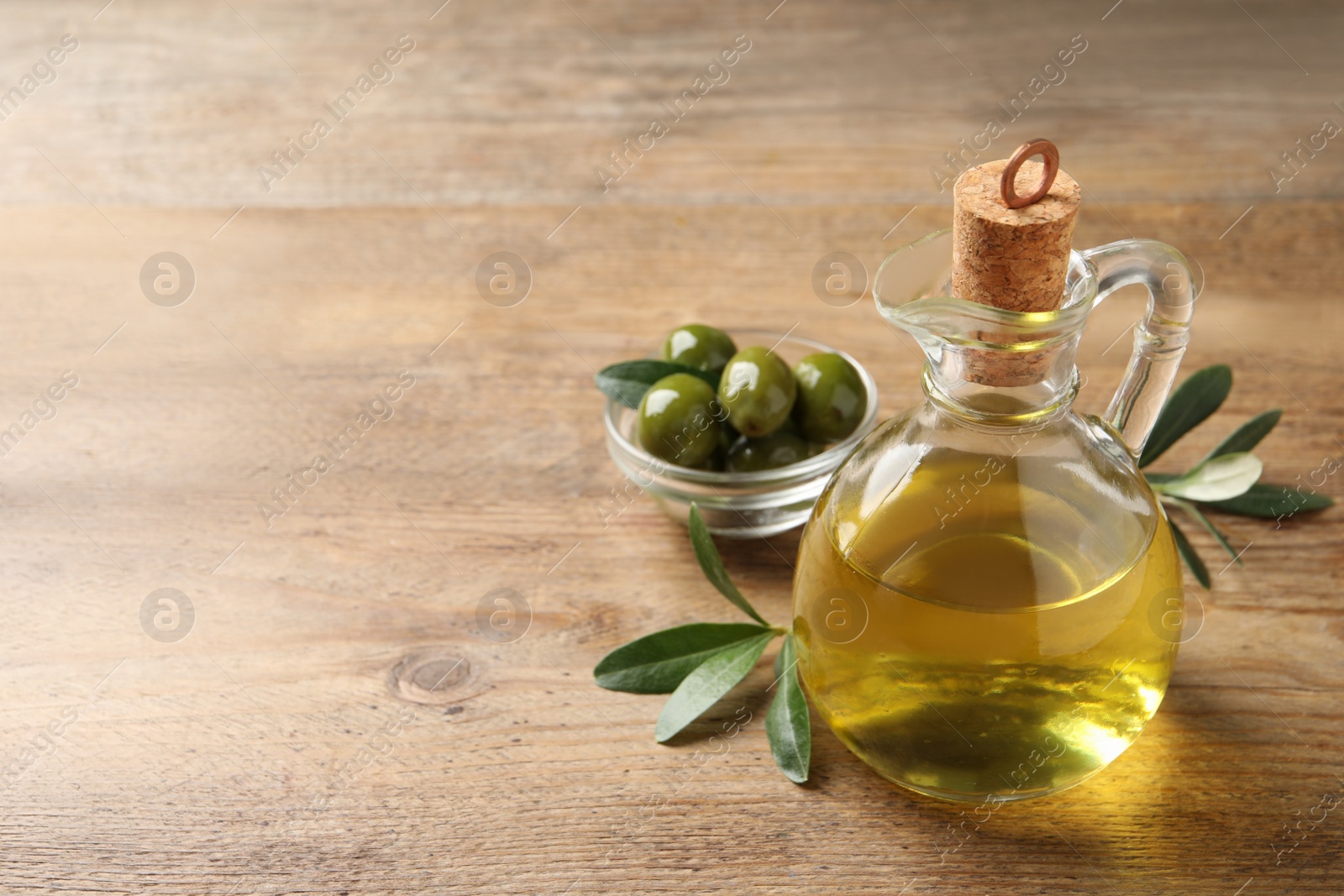 Photo of Glass jug of oil, ripe olives and green leaves on wooden table. Space for text