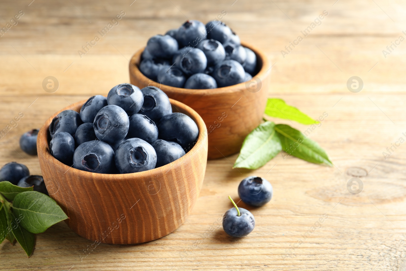 Photo of Bowls of fresh tasty blueberries and leaves on wooden table, space for text