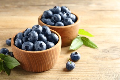 Bowls of fresh tasty blueberries and leaves on wooden table, space for text