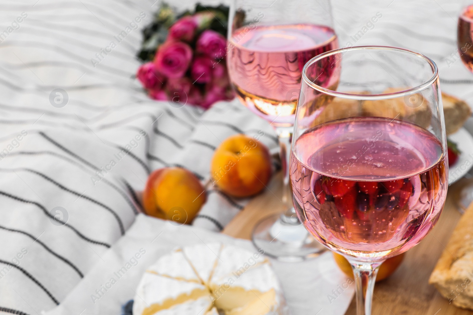 Photo of Glasses of delicious rose wine, flowers and food on white picnic blanket, closeup