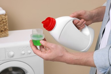 Photo of Man pouring fabric softener from bottle into cap near washing machine indoors, closeup