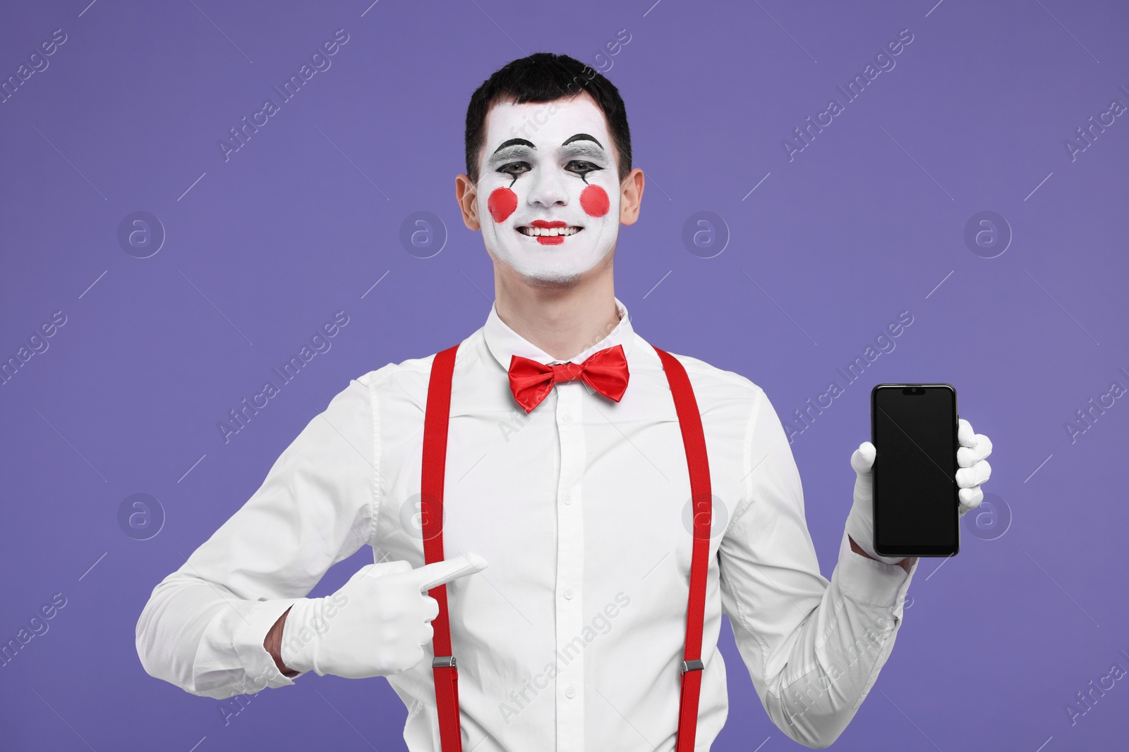 Photo of Funny mime artist pointing at smartphone on purple background