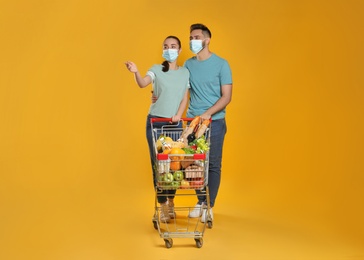 Couple with protective masks and shopping cart full of groceries on yellow background