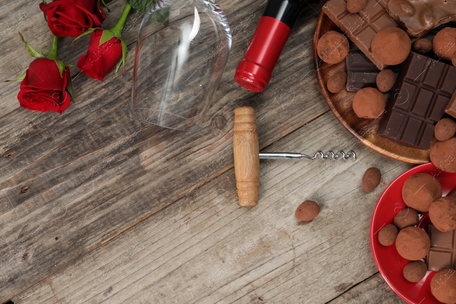 Photo of Bottle of red wine, glass, chocolate sweets, corkscrew and roses on wooden table, flat lay. Space for text