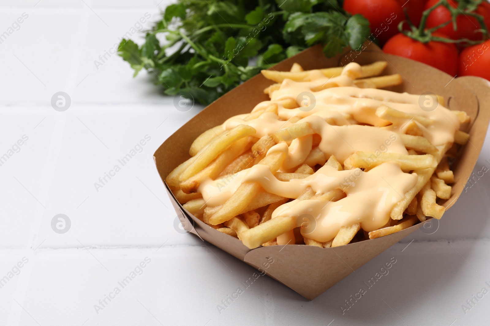 Photo of Delicious French fries with cheese sauce, tomatoes and parsley on white tiled table, closeup. Space for text