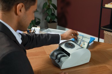 Photo of Man putting money into banknote counter at wooden table indoors