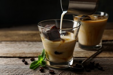 Photo of Pouring milk into glass of delicious iced coffee, mint and beans on wooden table