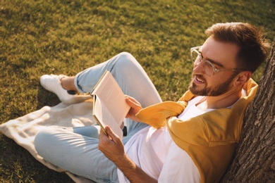 Young man reading book on green grass near tree in park