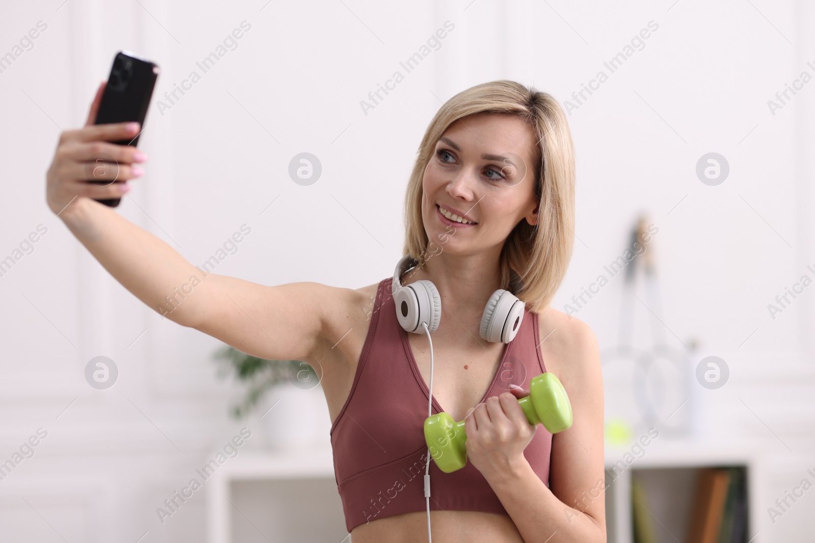 Photo of Smiling sports blogger holding dumbbell while streaming online fitness lesson with smartphone at home