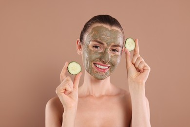 Photo of Beautiful woman with clay mask holding pieces of cucumber on light brown background