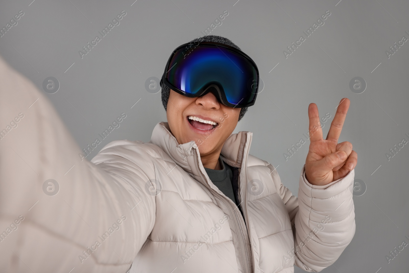 Photo of Winter sports. Happy man in ski goggles showing V-sign while taking selfie on grey background