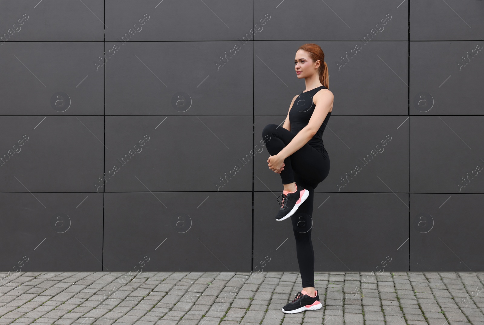 Photo of Beautiful woman in gym clothes doing exercises near dark grey wall on street, space for text