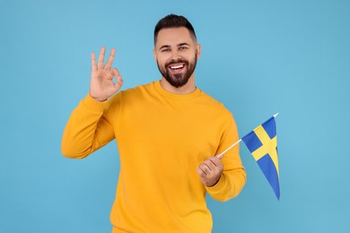 Photo of Young man with flag of Sweden showing ok gesture on light blue background