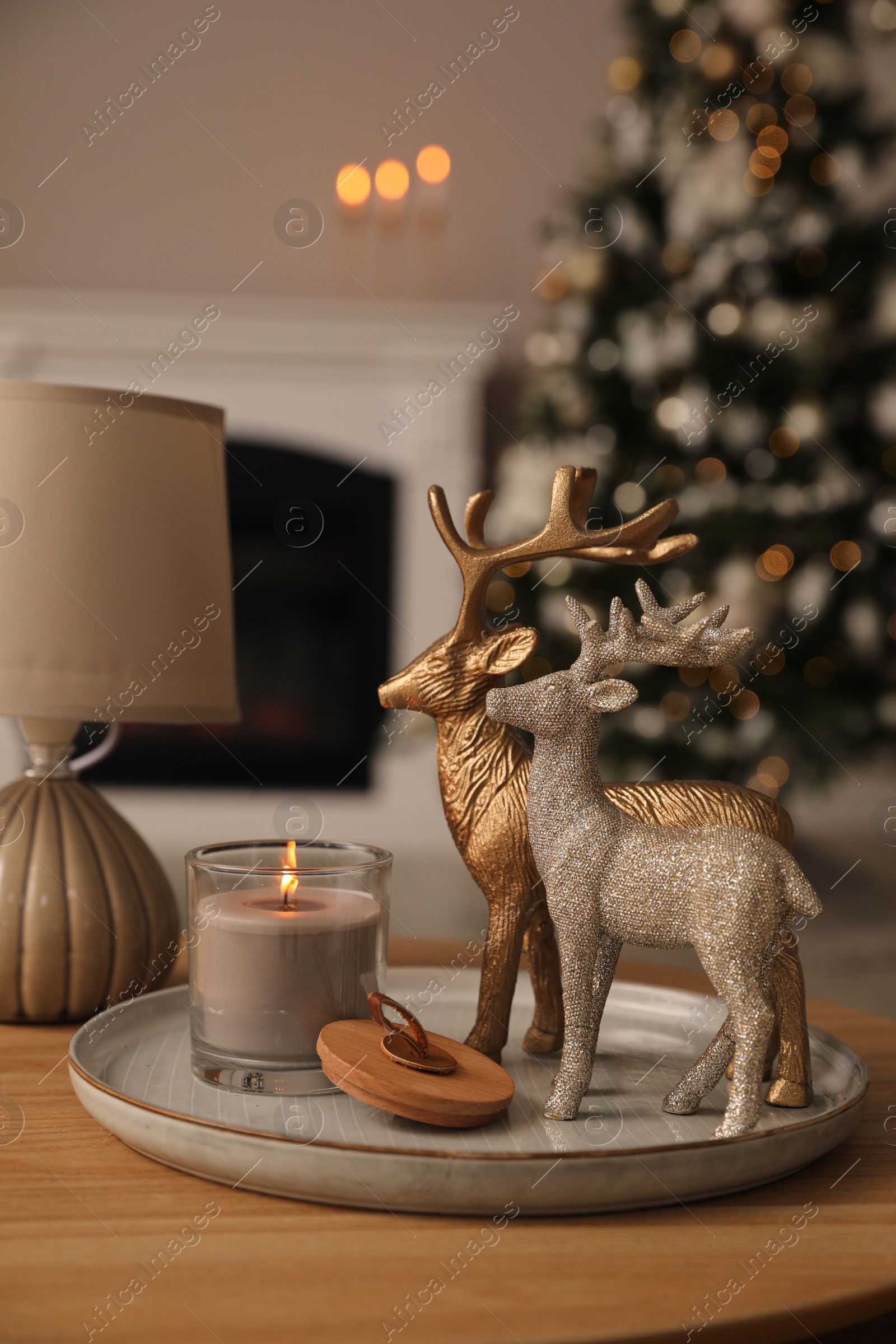 Photo of Deer figures, burning candle with lamp on wooden table and Christmas tree in living room