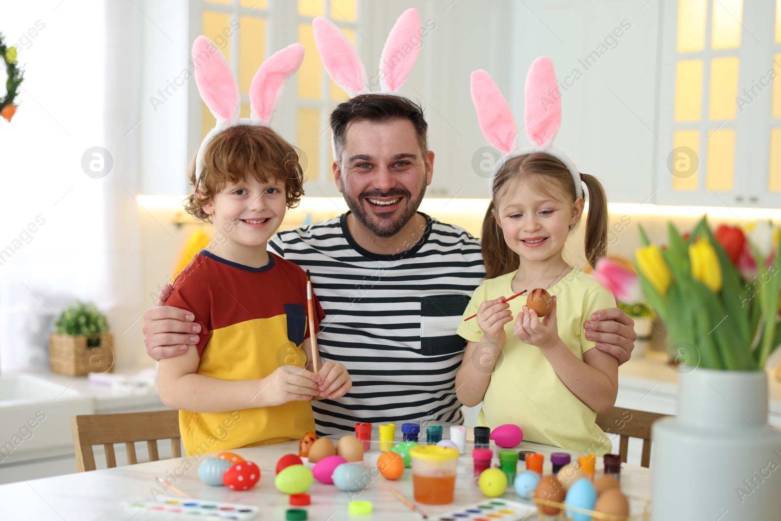 Photo of Painting Easter eggs. Happy father and his cute children with bunny ears at white marble table in kitchen