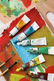 Photo of Tubes of colorful oil paints, brushes and canvas with abstract painting on white table, flat lay