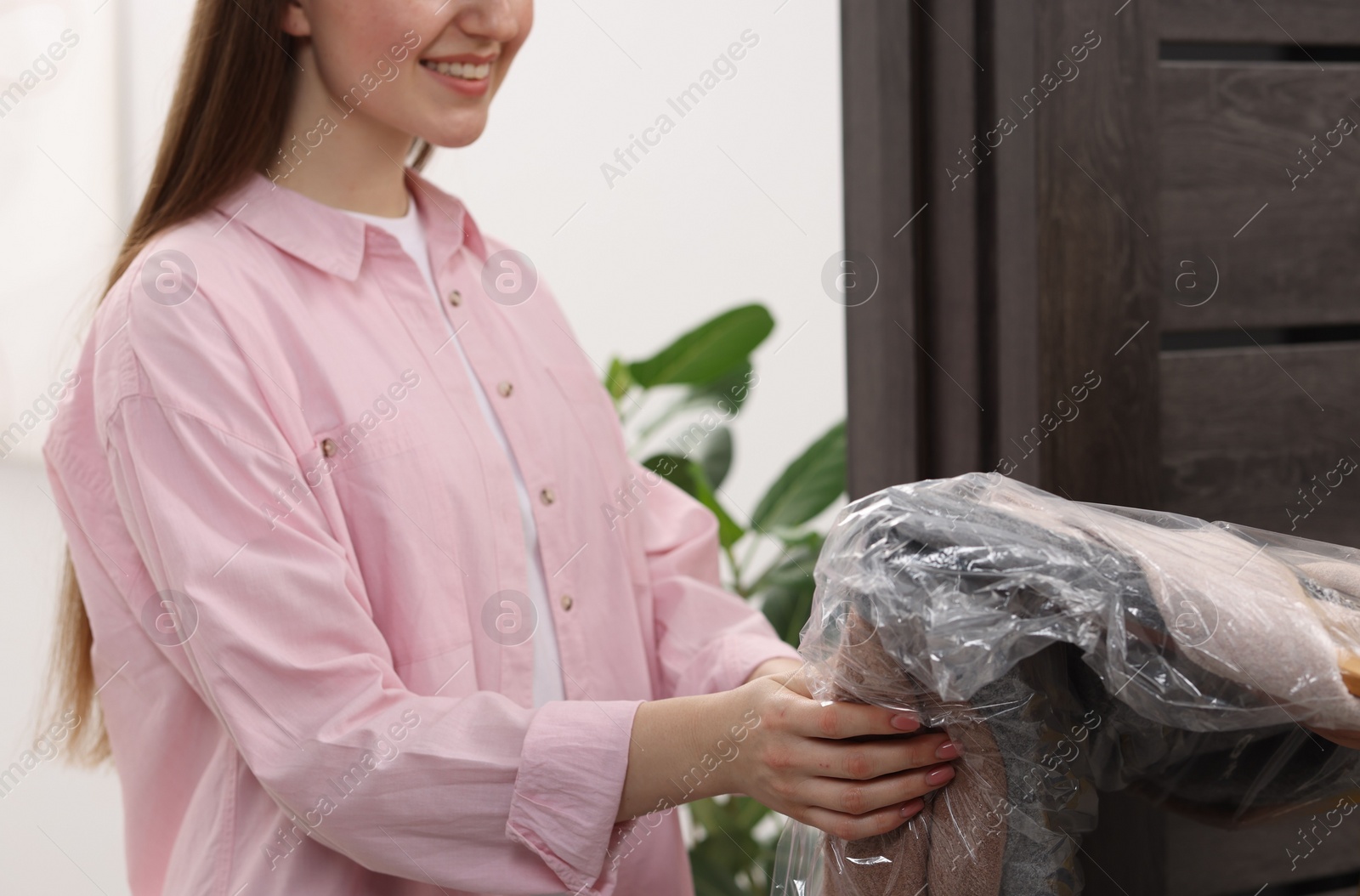 Photo of Dry-cleaning delivery. Happy woman holding coat in plastic bag indoors, closeup