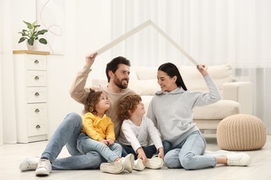Photo of Family housing concept. Happy woman and her husband holding plastic roof while sitting with kids on floor at home