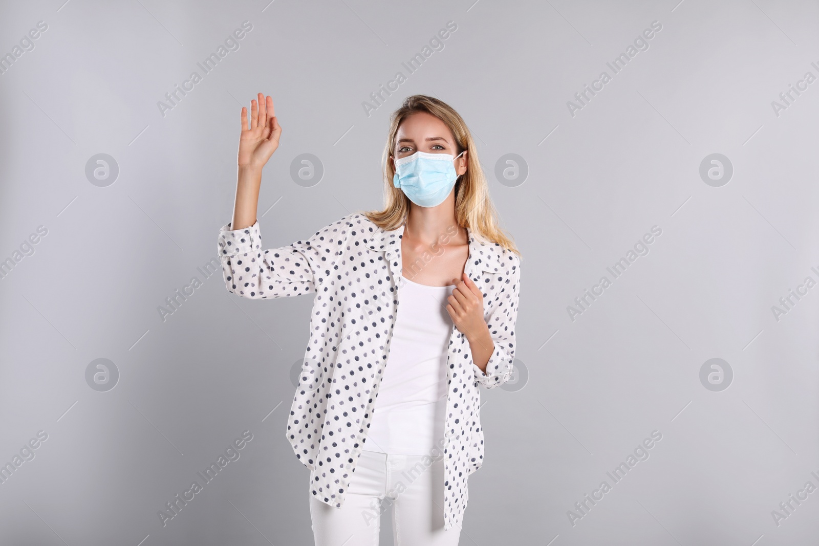 Photo of Woman in protective face mask showing hello gesture on grey background. Keeping social distance during coronavirus pandemic