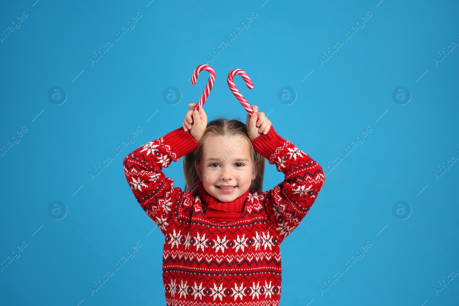 Photo of Cute little girl in knitted Christmas sweater holding candy canes on blue background