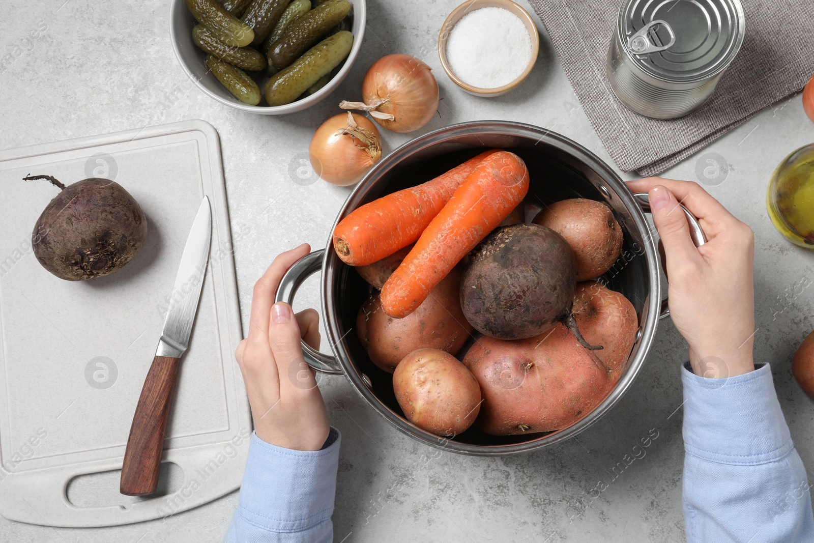 Photo of Woman holding pot with fresh vegetables at white table, top view. Cooking vinaigrette salad