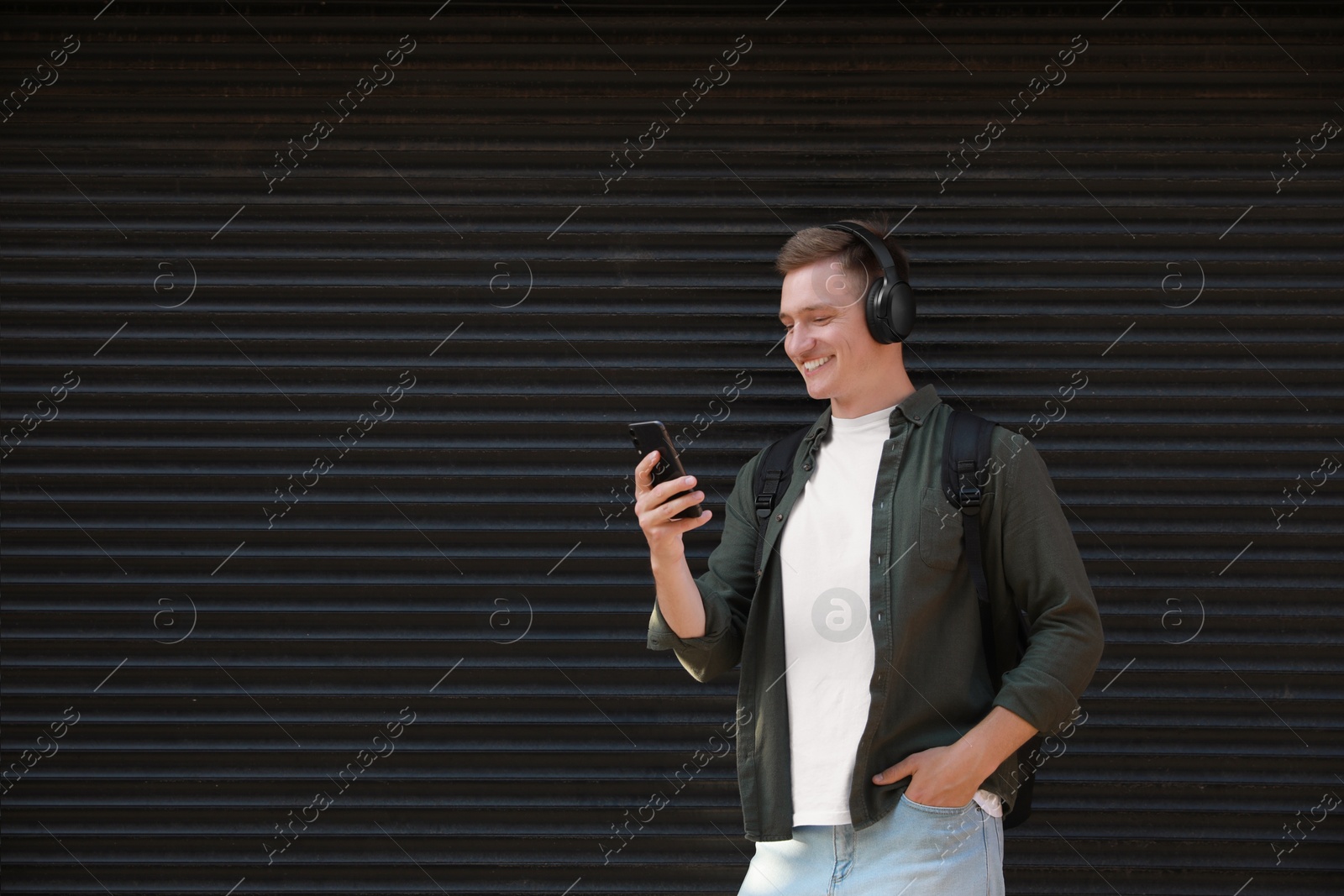 Photo of Smiling man in headphones using smartphone near shutters outdoors. Space for text