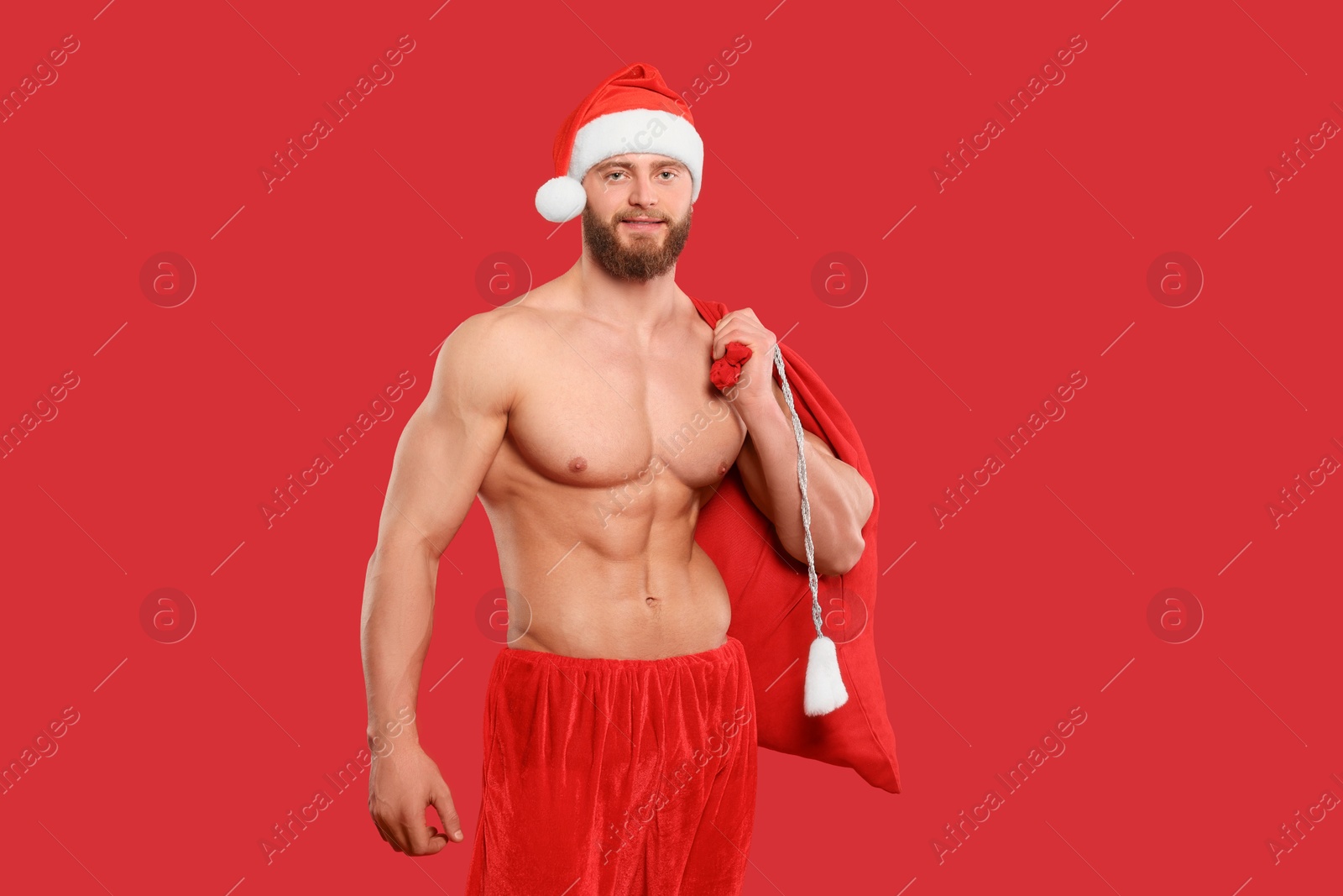 Photo of Muscular young man in Santa hat holding bag with presents on red background