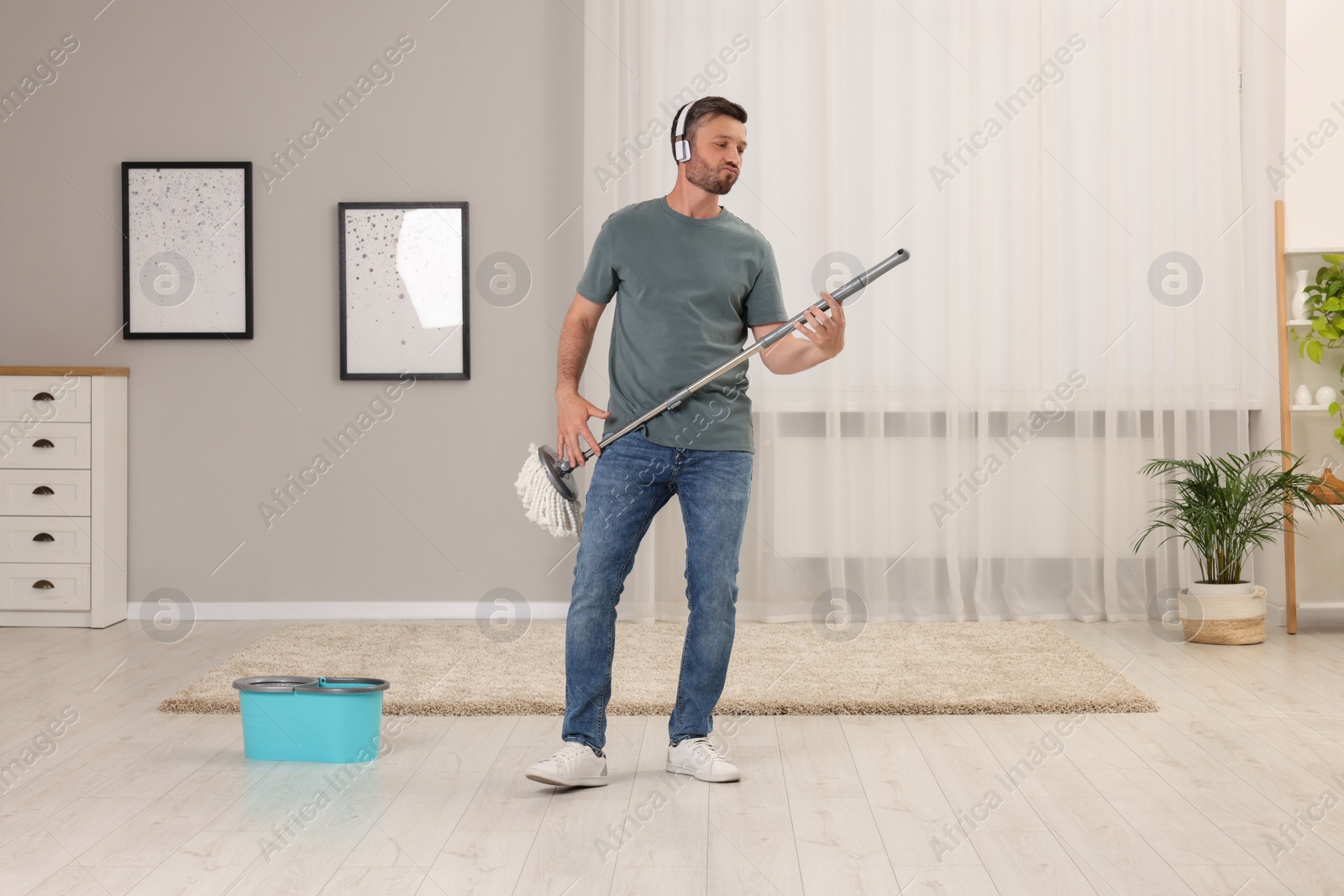 Photo of Enjoying cleaning. Happy man in headphones with mop at home