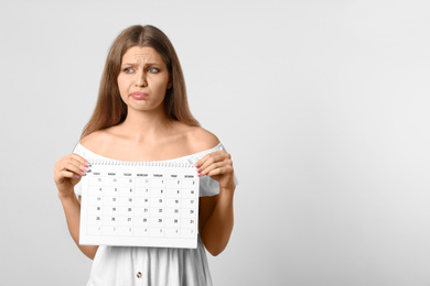 Emotional young woman holding calendar with marked menstrual cycle days on light background. Space for text