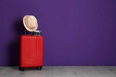 Travel suitcase with hat and camera on wooden floor near purple wall, space for text. Summer vacation