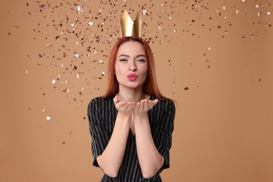 Photo of Beautiful young woman with princess crown blowing kiss under falling confetti on beige background