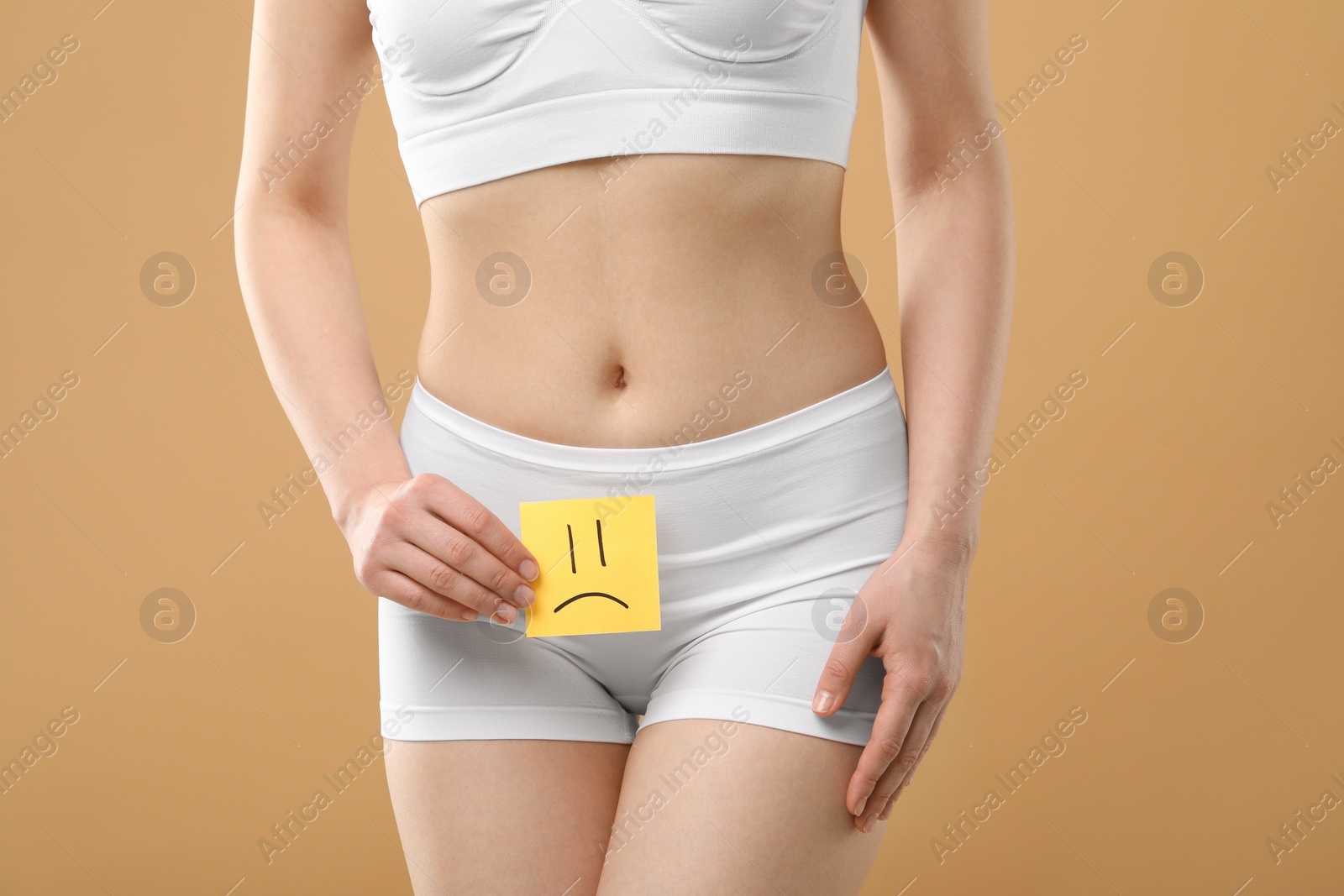 Photo of Cystitis. Woman holding sticky note with drawn sad face on beige background, closeup