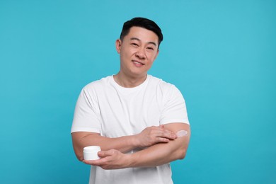 Photo of Handsome man applying body cream onto his arm on light blue background