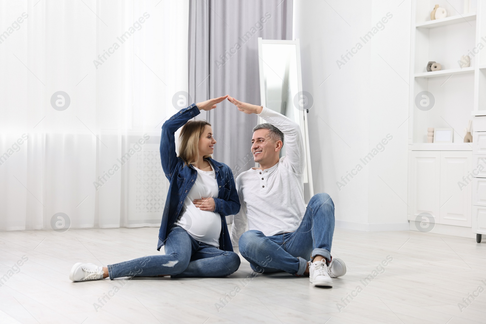 Photo of Young family housing concept. Pregnant woman with her husband forming roof with their hands while sitting on floor at home