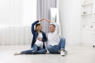 Young family housing concept. Pregnant woman with her husband forming roof with their hands while sitting on floor at home