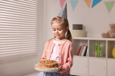 Birthday celebration. Cute little girl in party hat holding tasty cake with burning candles at home