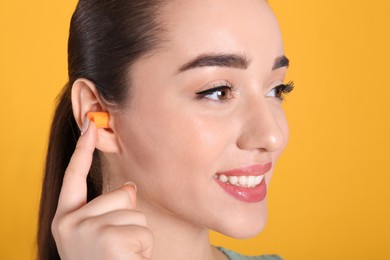 Young woman inserting foam ear plug on yellow background, closeup