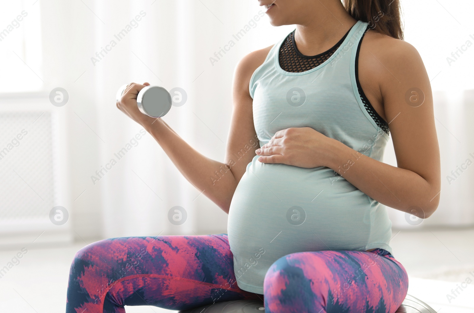 Photo of Young pregnant woman in fitness clothes lifting dumbbell at home, closeup